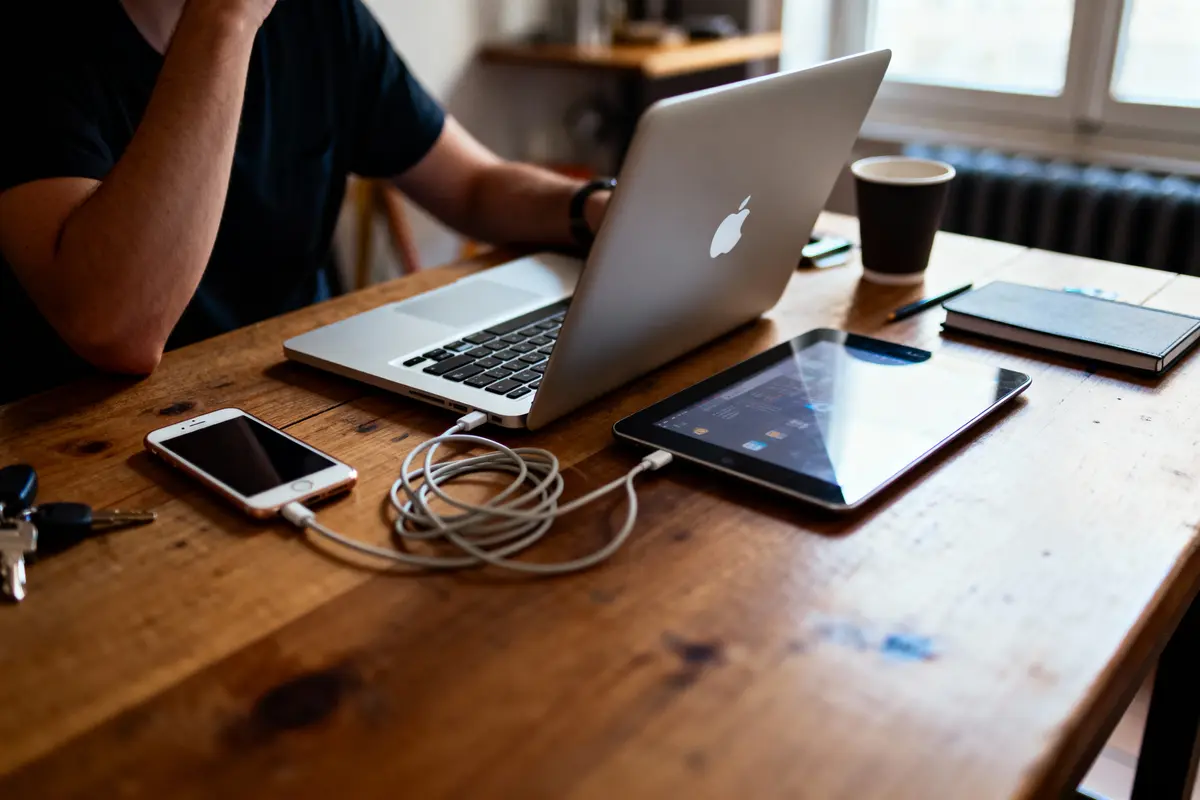 Personne travaillant sur un MacBook avec smartphone, tablette, café et carnet sur une table en bois dans un espace lumineux.