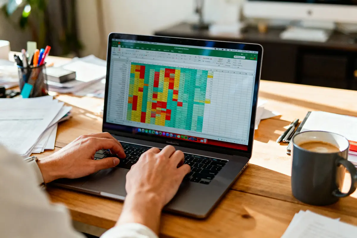 Personne travaillant sur un tableur Excel coloré avec tableau de données sur ordinateur portable, bureau en bois avec café.