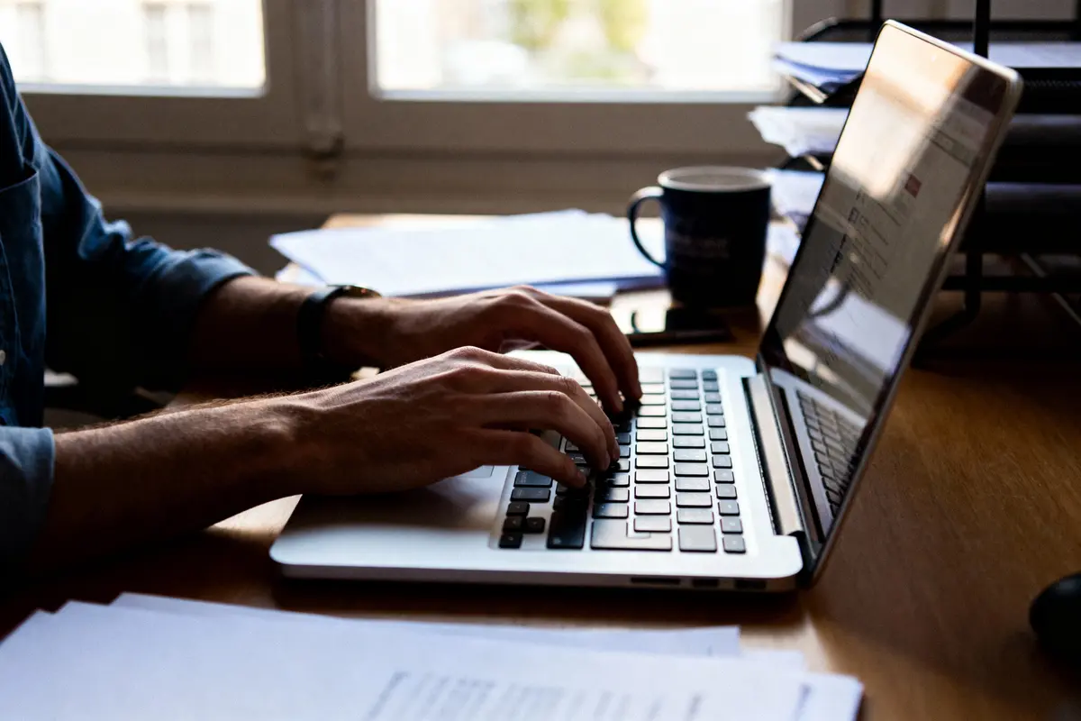 Personne tapant sur un ordinateur portable sur un bureau en bois avec tasse de café et documents en arrière-plan.