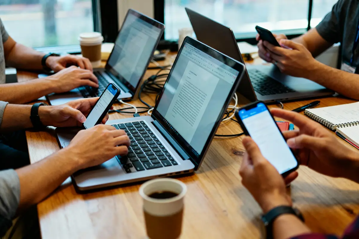 Équipe collaborative travaillant sur ordinateurs portables et smartphones autour d'une table en bois avec cafés dans un espace de coworking moderne.