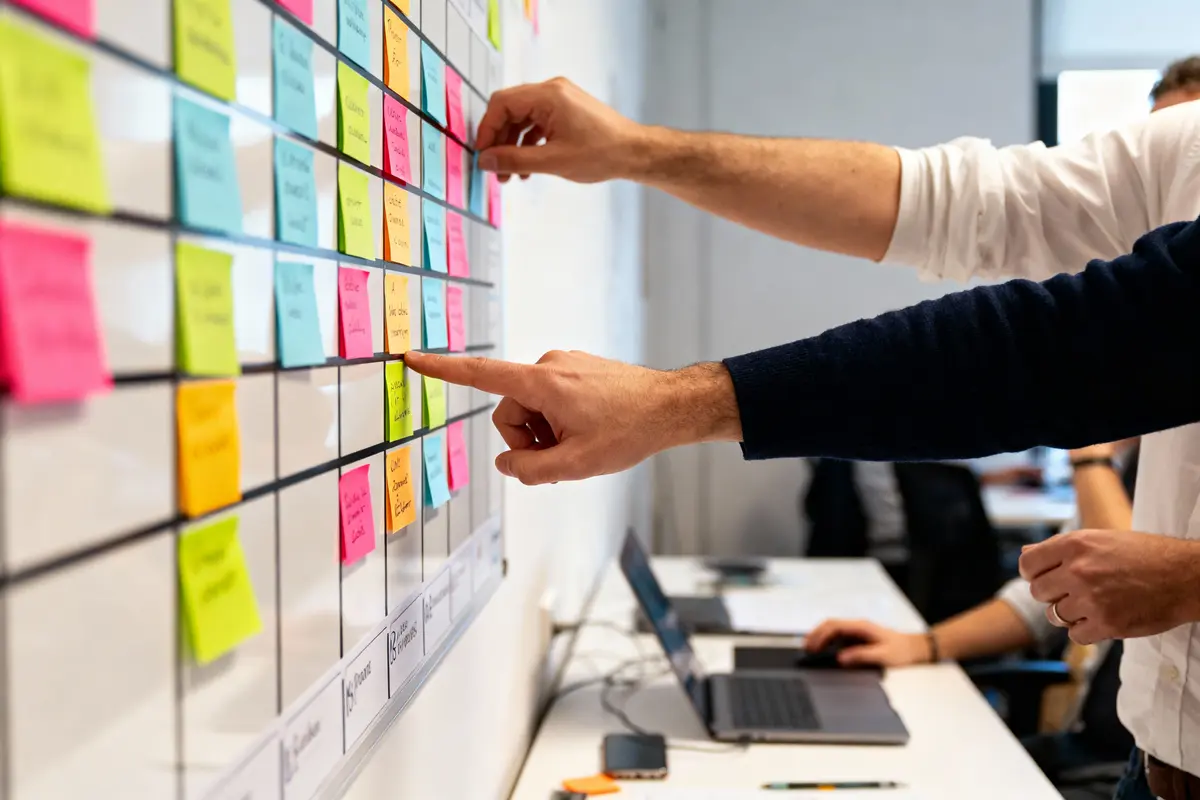 Deux personnes pointent des post-it colorés sur un tableau de planification lors d'une réunion de travail collaboratif en bureau.